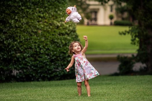 A young girl joyfully throws her doll in the air while playing outdoors on a summer day.