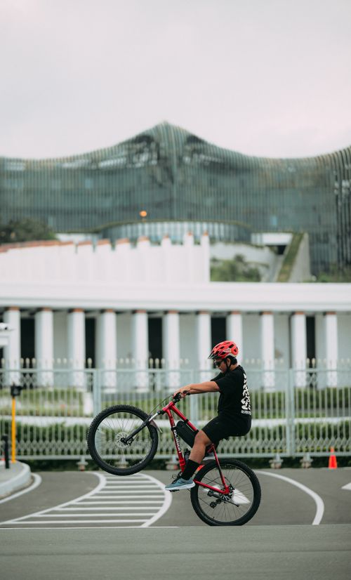 Free A cyclist in a red helmet performs a wheelie on a bicycle in an urban setting with unique architecture in the background. Stock Photo