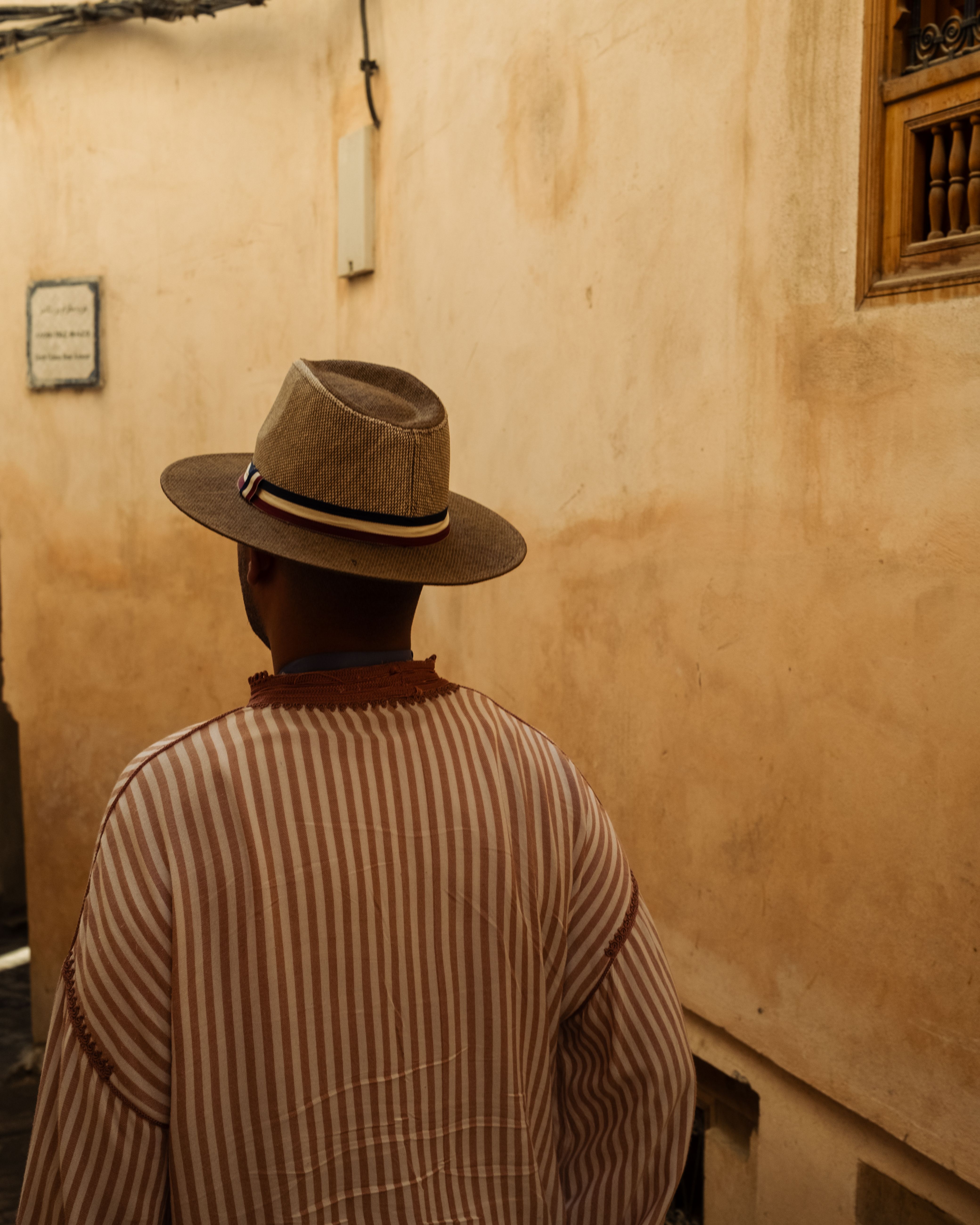 A person in a striped garment and hat walks through a sunlit narrow alleyway in a historic city.