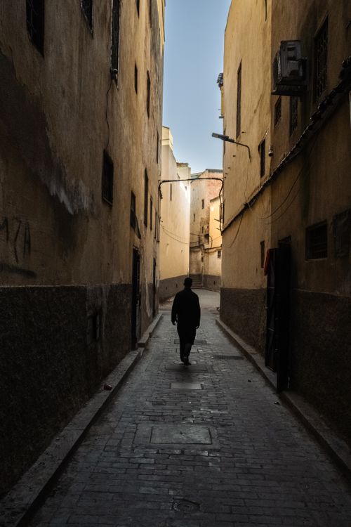 Free A lone silhouette walks through a narrow alleyway between aged buildings at dusk. Stock Photo