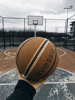 A detailed view of a basketball held over an outdoor court with hoop in focus.