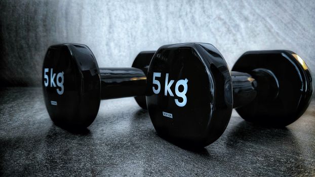 Close-up of black 5kg dumbbells on a textured dark surface, highlighting fitness and strength training equipment.