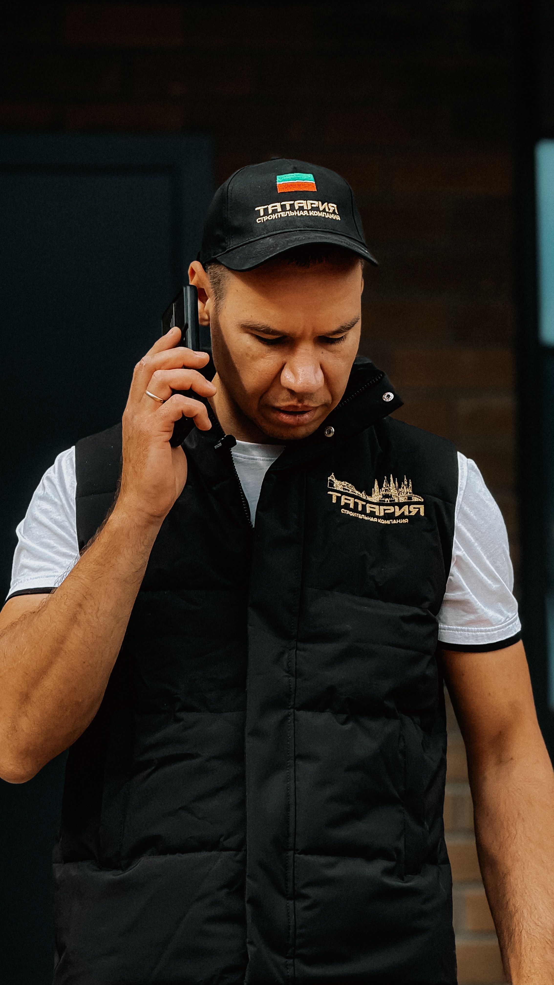 Casual man wearing black vest and cap speaking on smartphone outdoors.