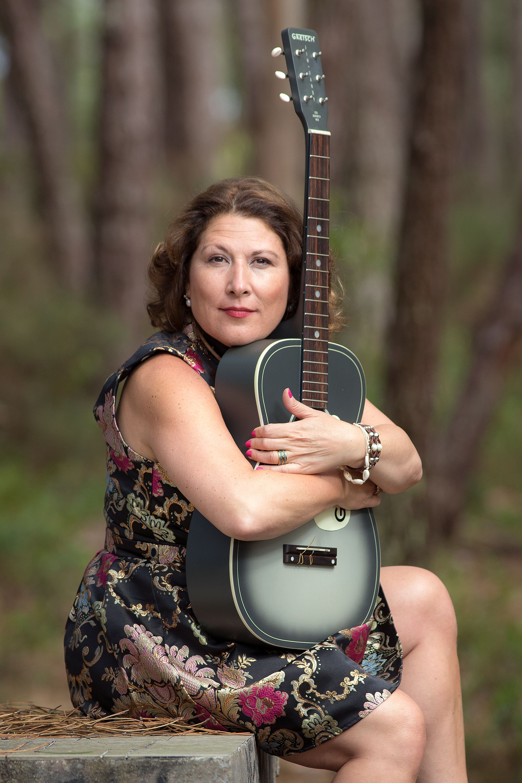 Woman in floral dress holds a guitar in a serene outdoor forest setting.