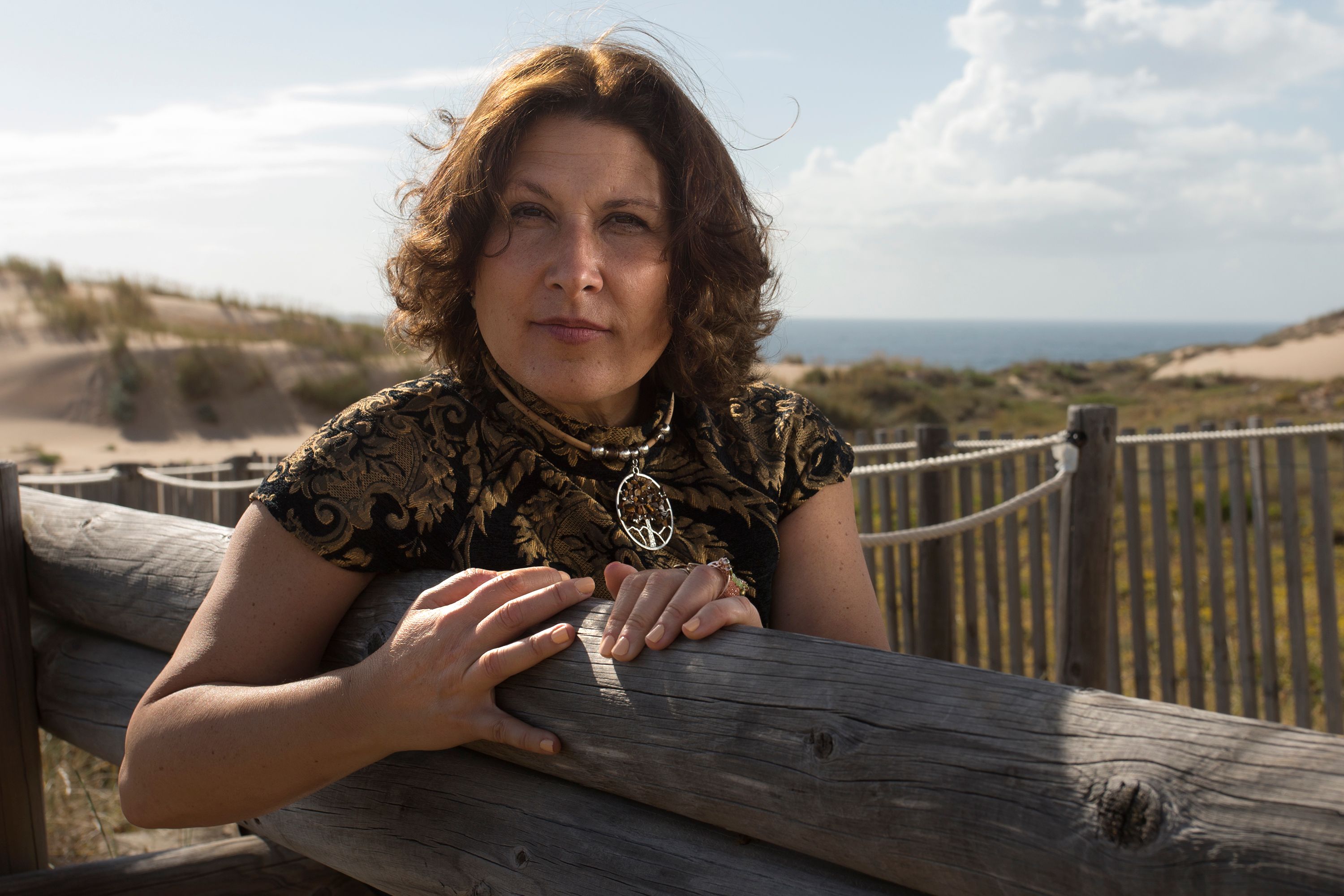 A woman leans against a wooden fence with beautiful dunes and ocean in the background.