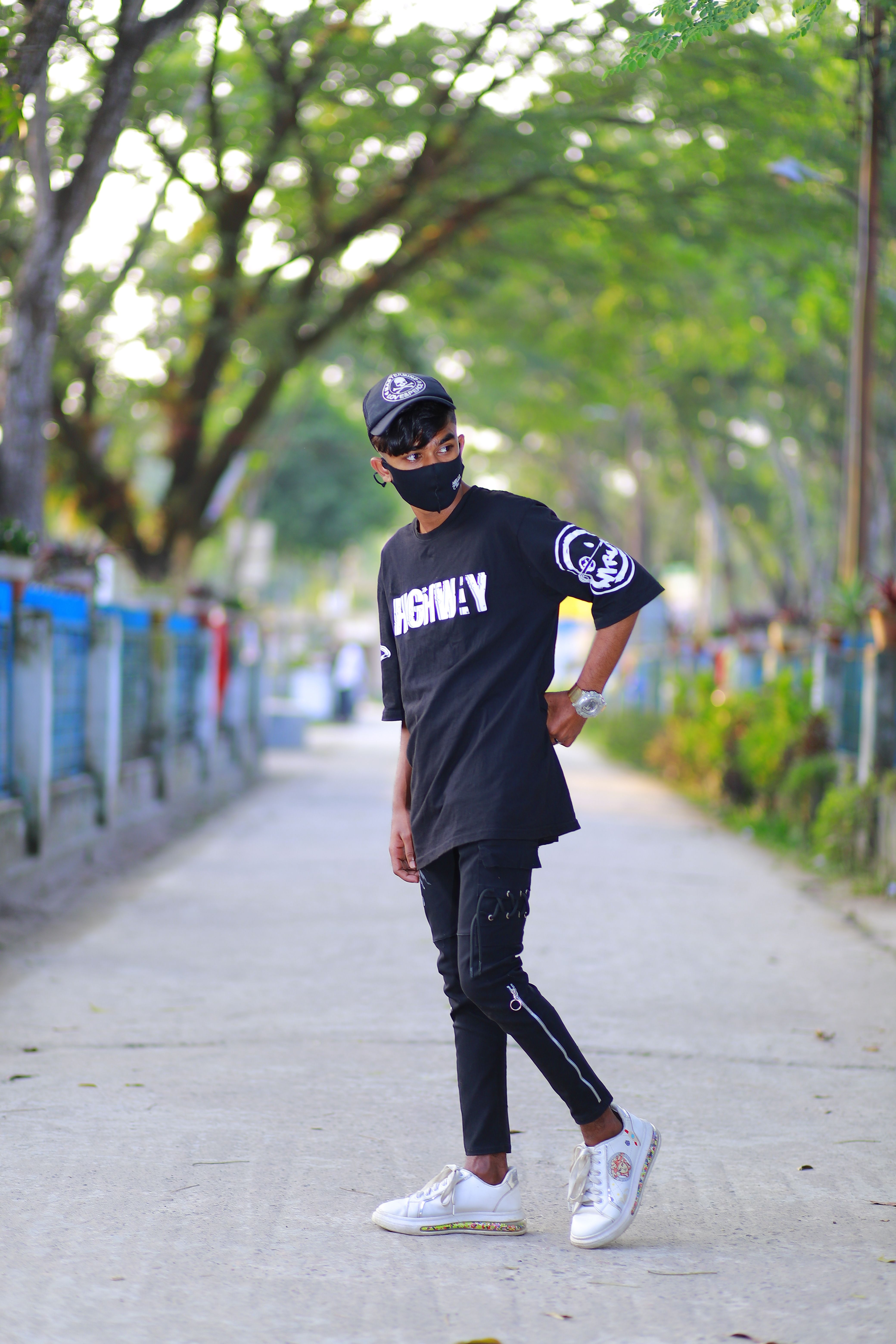 A fashionable young man wearing streetwear, standing confidently on a tree-lined urban pathway.