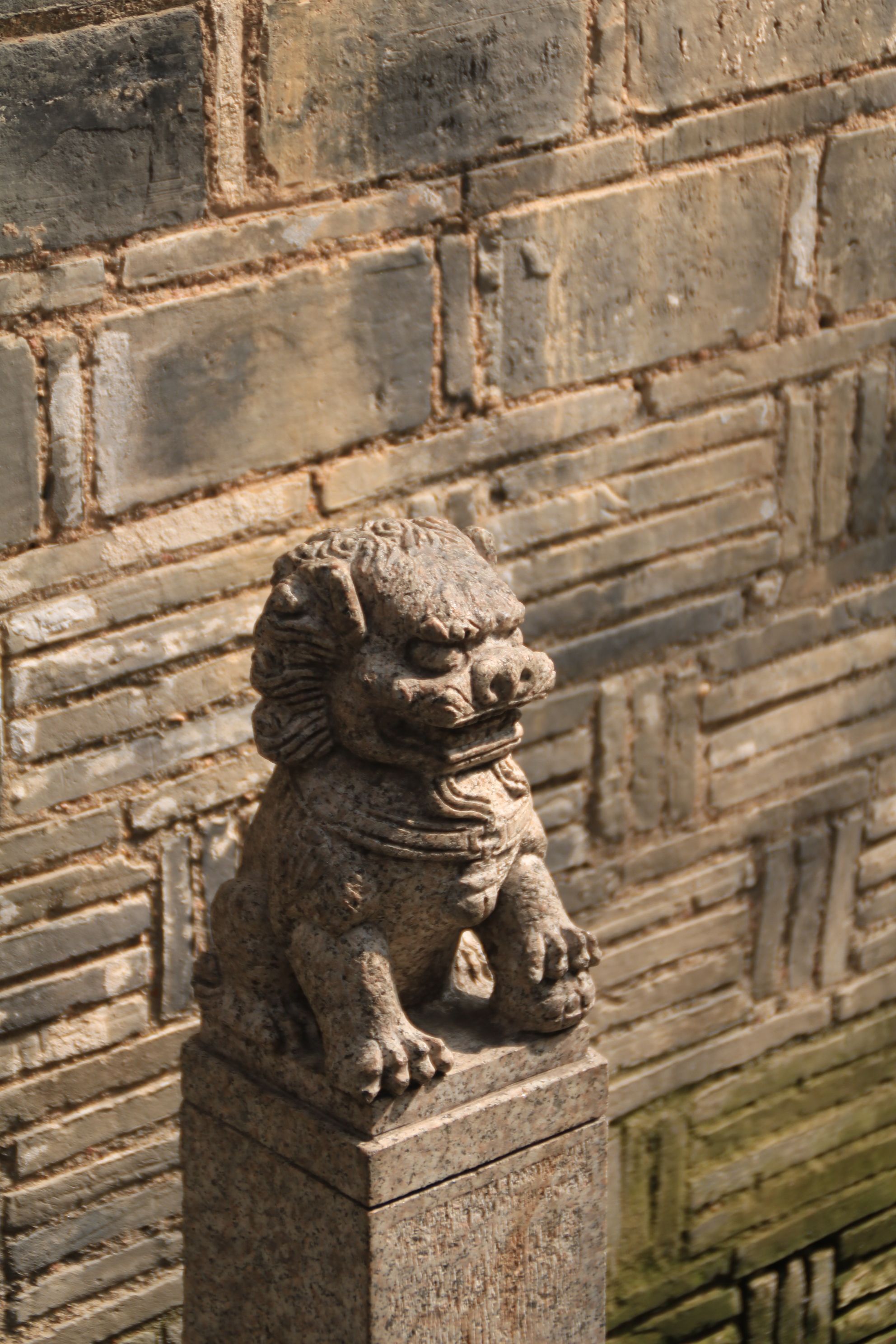 Gratuit Une statue de chien Fô en pierre se dresse en faction devant un mur de briques rustique, évoquant une culture ancienne. Photos