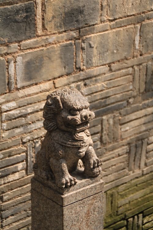 Gratuit Une statue de chien Fô en pierre se dresse en faction devant un mur de briques rustique, évoquant une culture ancienne. Photos