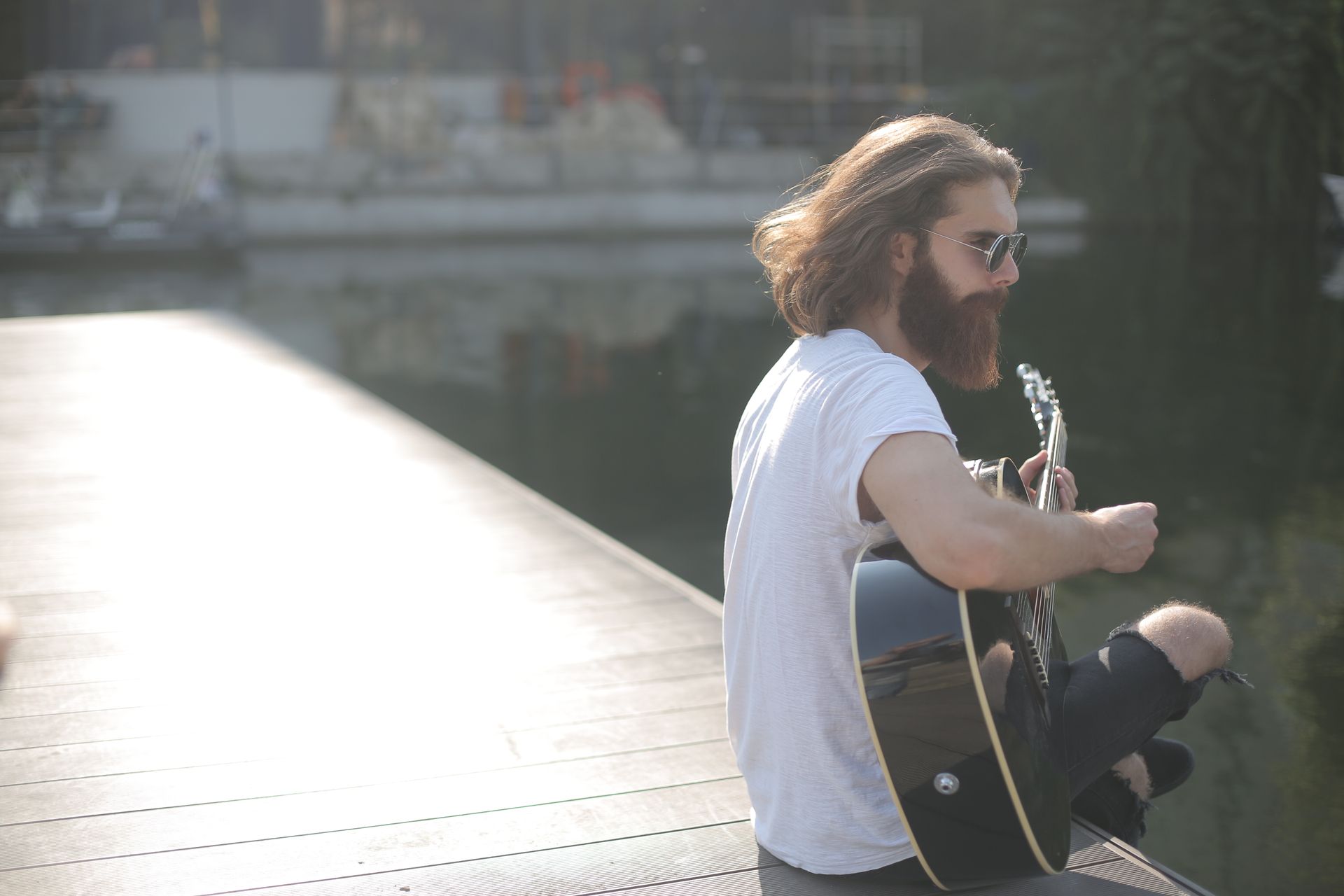 A bearded man sits on a dock playing an acoustic guitar under the daylight.