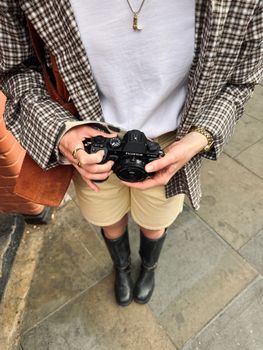 Close-up of a person holding a camera, standing on a London sidewalk. Fashionable attire and urban setting.