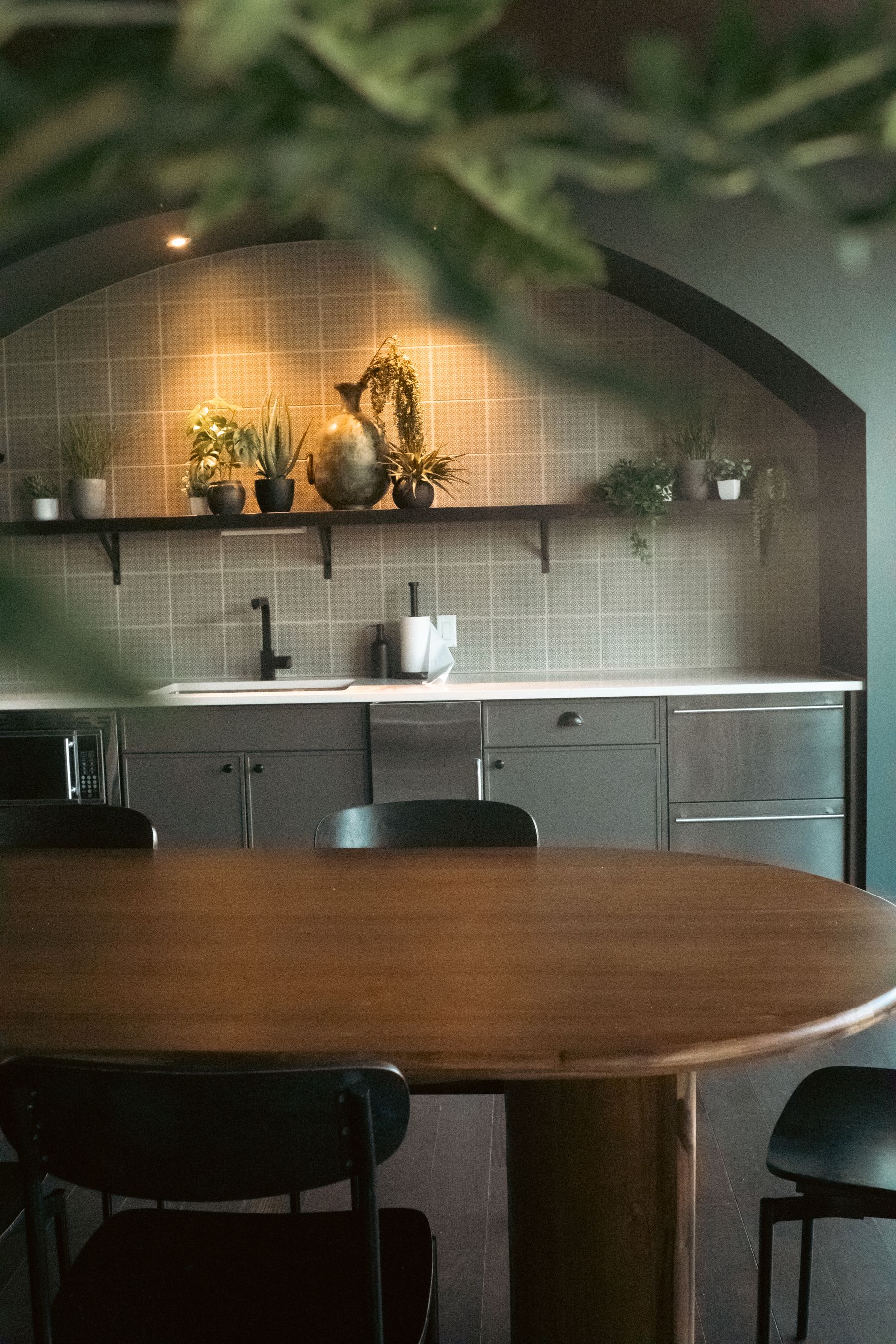 Dark kitchen cabinetry below a tiled arch and planted shelf, with a rounded wooden table