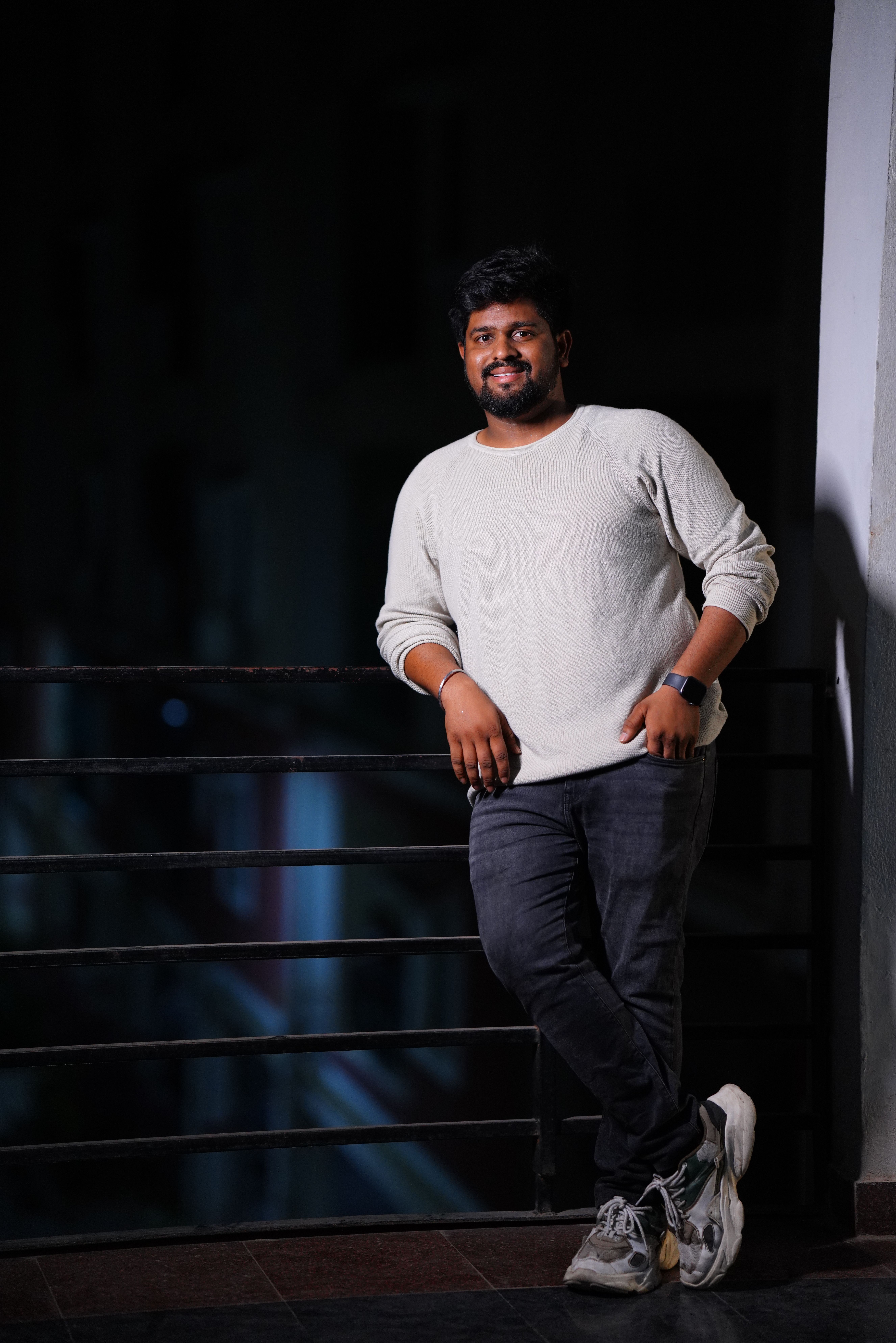 A smiling man in casual attire stands on a balcony at night, showcasing relaxed confidence.