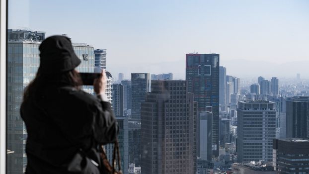 A person photographs the stunning Osaka skyline from a high-rise building window, showcasing urban architecture.