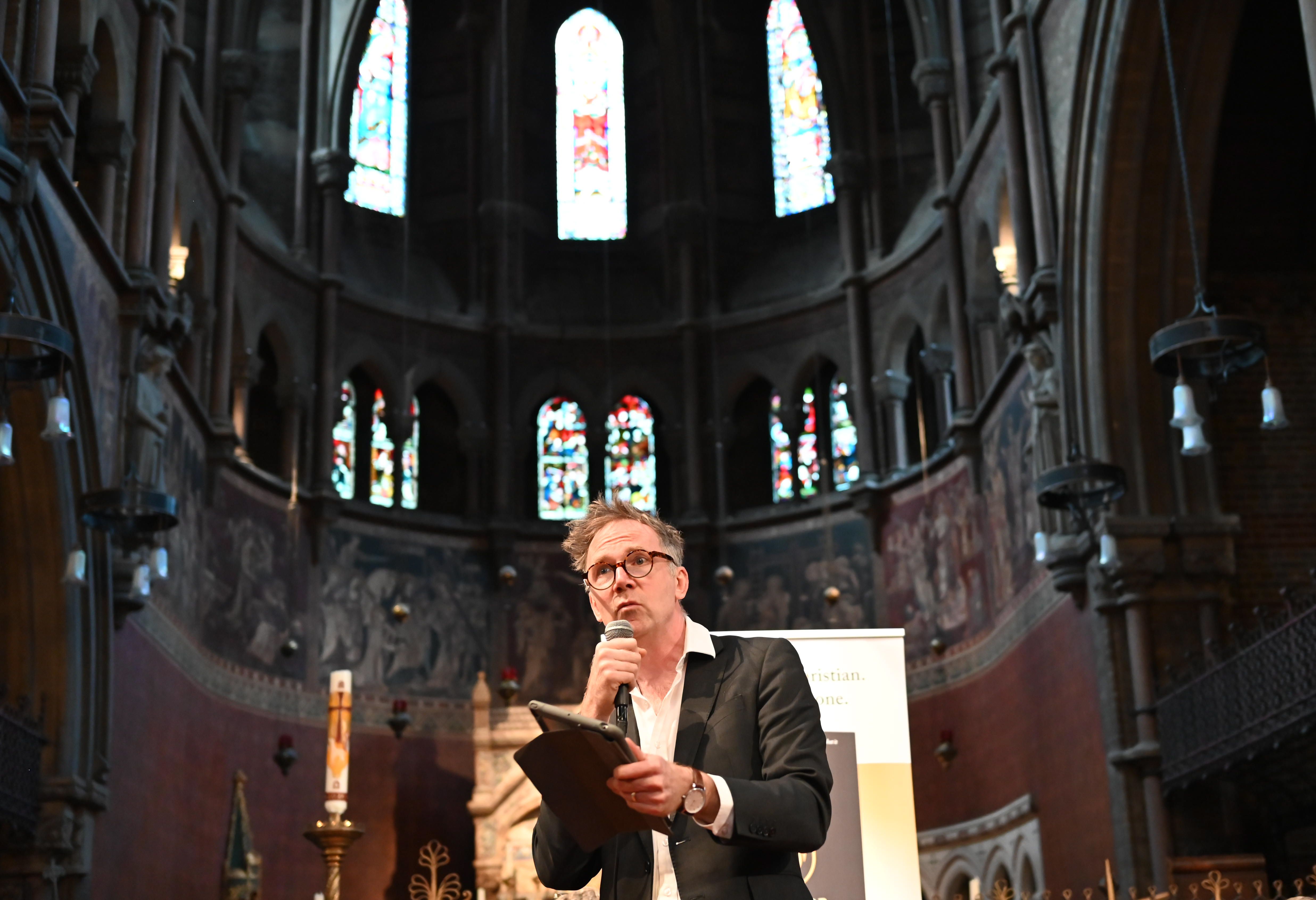 Man speaking in a historic church, surrounded by stained glass windows and ornate architecture.