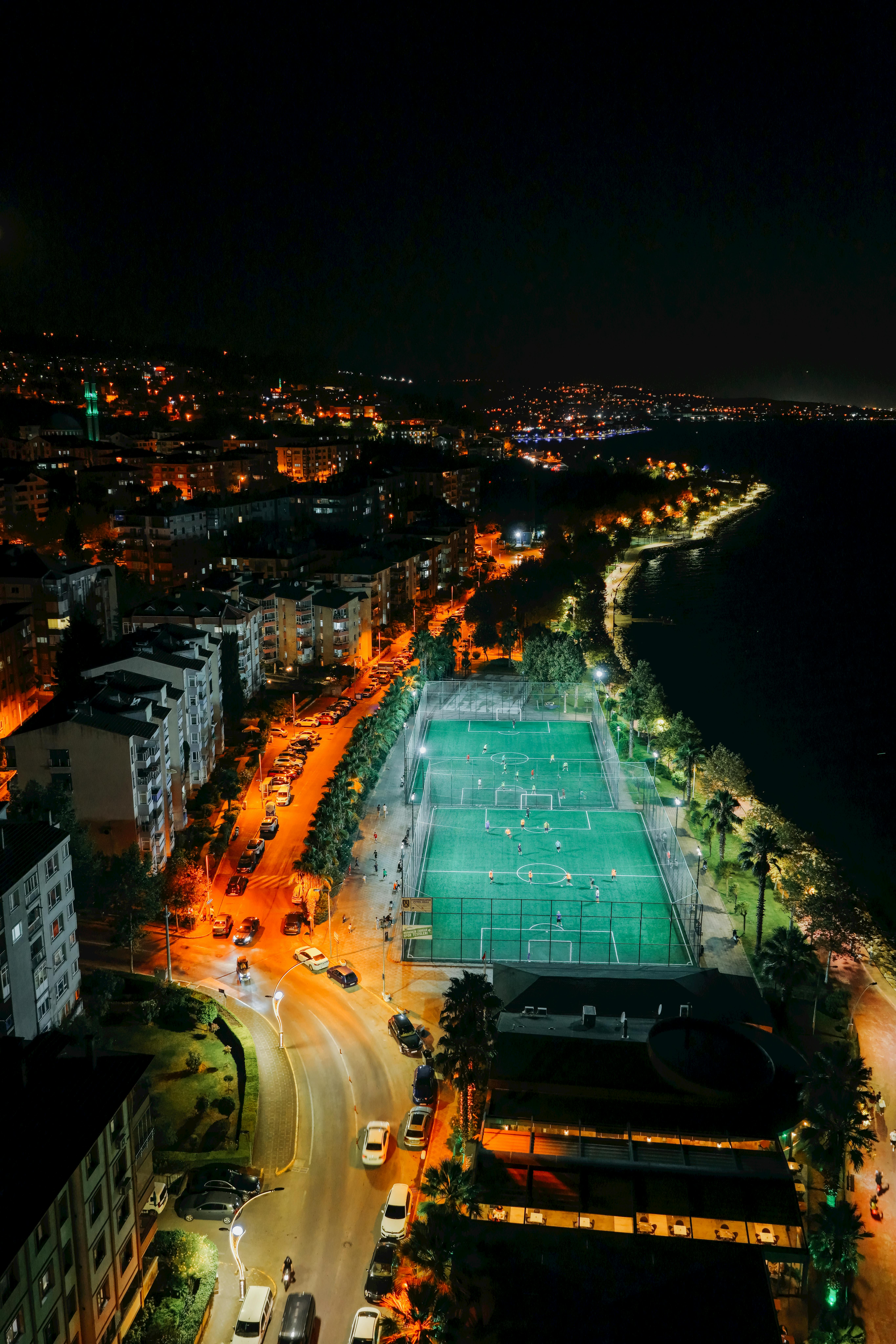 A vibrant aerial night view of a city by the coast featuring a lit soccer field and illuminated streets.