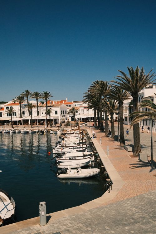 Free Picturesque view of a sunny harbor in Menorca with boats and palm-lined promenade. Stock Photo