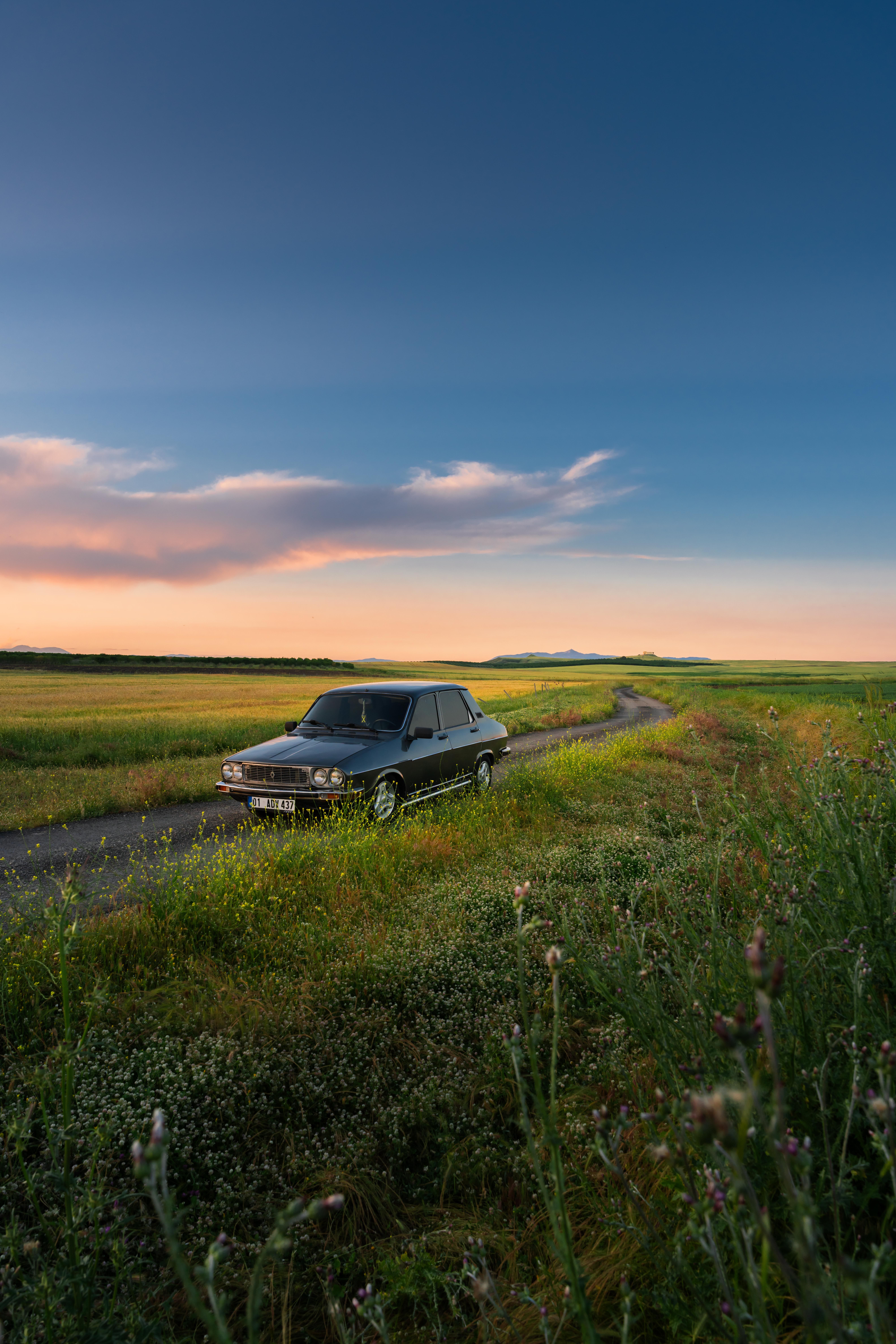 A vintage car parked along a picturesque country road during sunset, surrounded by lush fields.