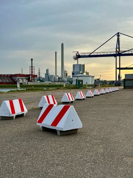 A row of red and white barriers at an industrial port with cranes and factories at sunset.
