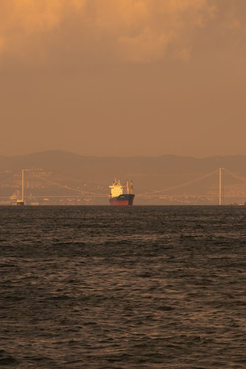 Free A cargo ship sails under an orange sunset with a bridge and mountains in the background. Stock Photo