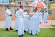 A priest performs a religious ceremony with acolytes in white robes outdoors.