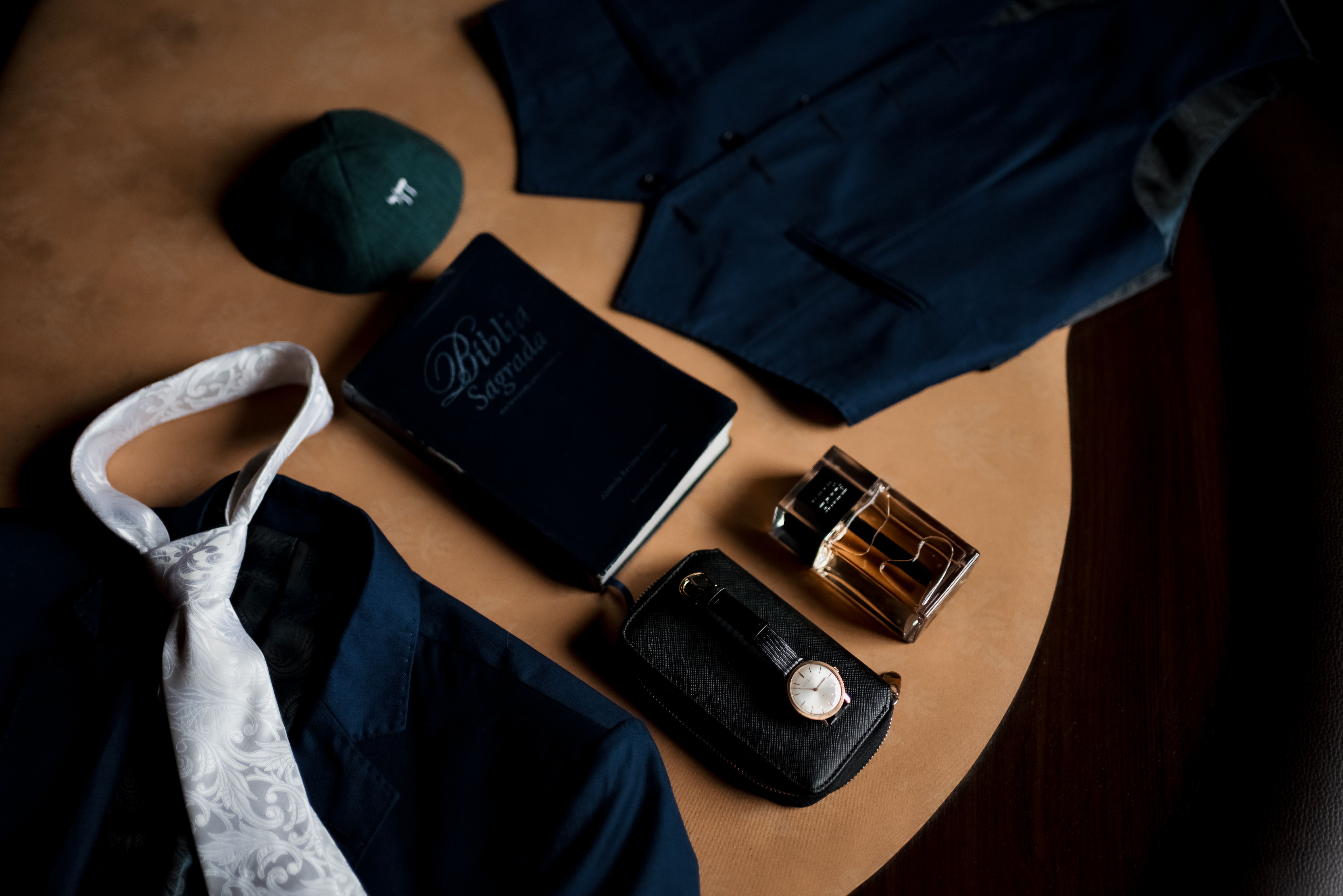 Flat lay of groom's wedding essentials including suit, tie, watch, and fragrance bottle.