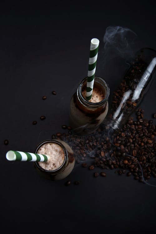 From above of similar glass jars with foamy cocoa cocktail and striped drinking tubes on black surface with grated chocolate