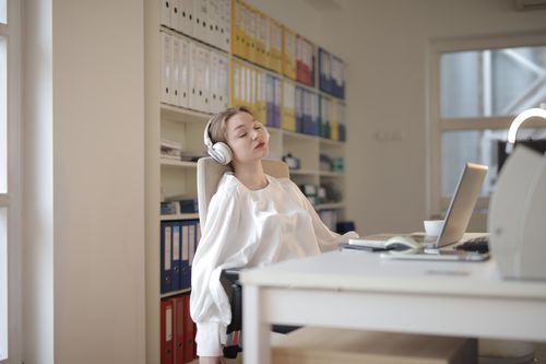 Free Woman in white shirt relaxing at office desk with headphones, appearing restful. Stock Photo