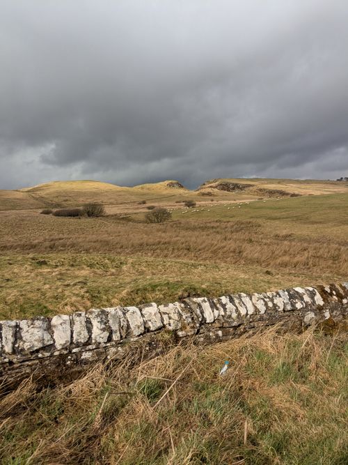 Free Dramatic stormy landscape featuring a stone wall and rolling grassy hills under dark clouds. Stock Photo