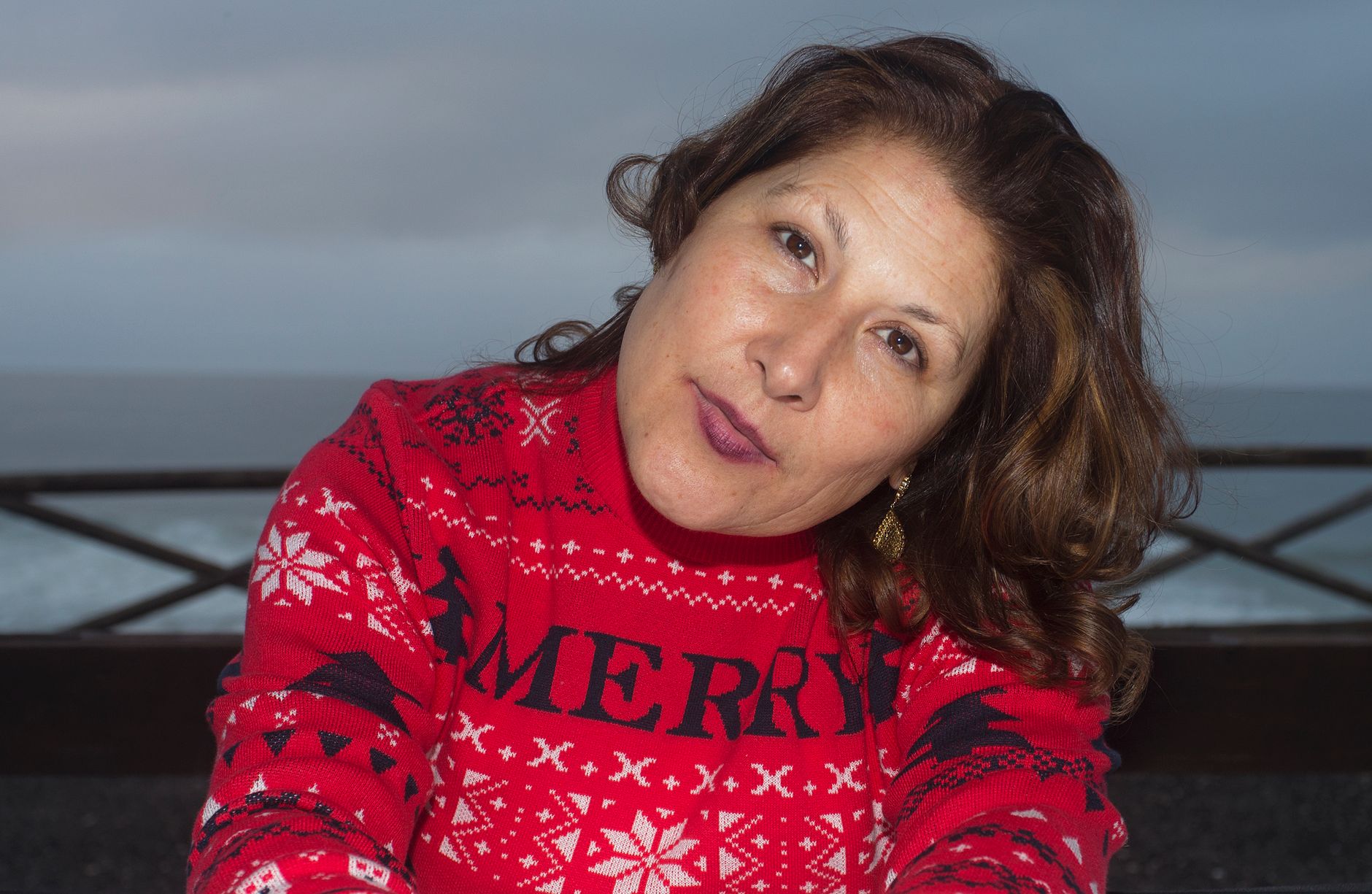 A woman in a red Christmas sweater poses by the sea on a cloudy day, showcasing holiday spirit.