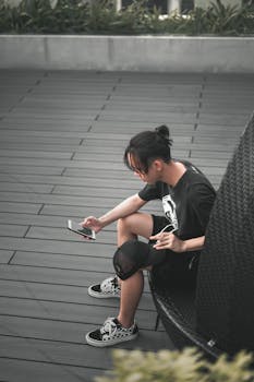 A young man in casual attire sits outside, engrossed in his smartphone, enjoying a relaxed summer day.