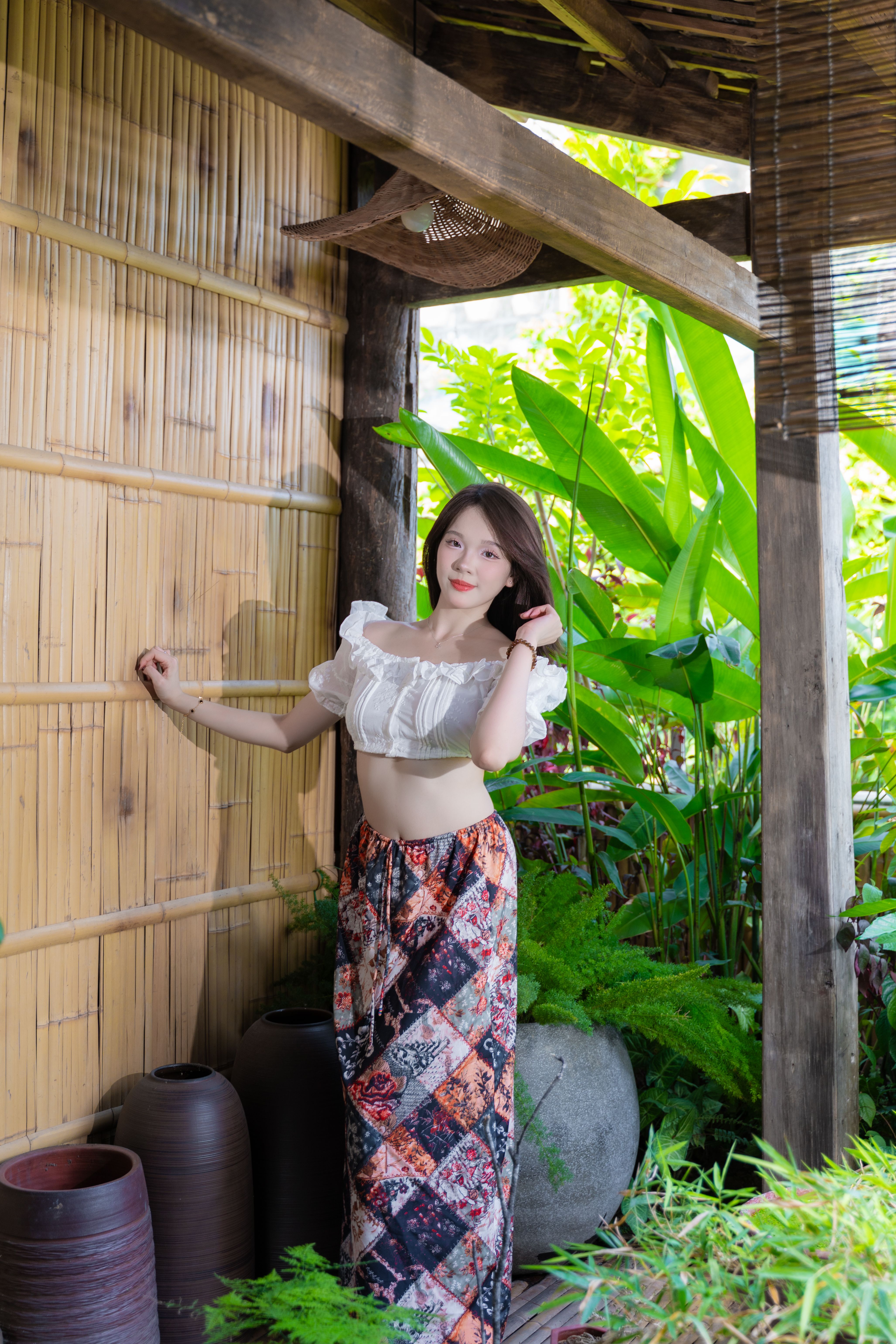 A woman stands in a tropical garden, wearing traditional attire with a bamboo backdrop.