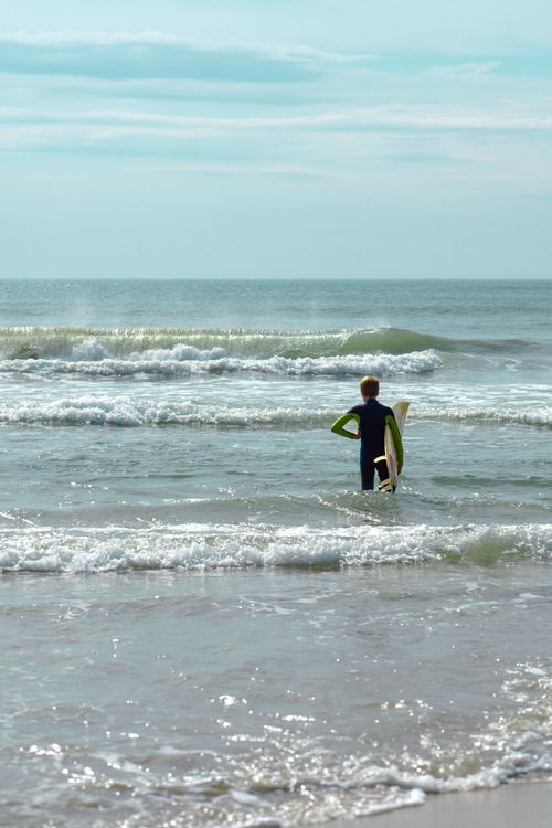 Free A boy stands in the ocean holding a surfboard, ready to surf under a bright blue sky. Stock Photo