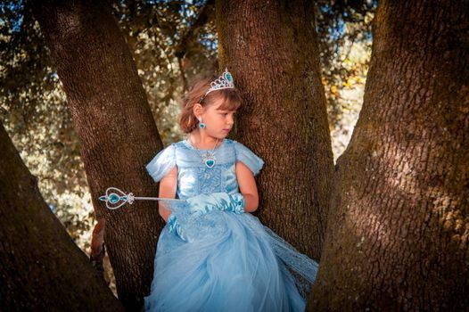 Young girl in a blue princess dress, tiara, and wand sitting in a tree outdoors.