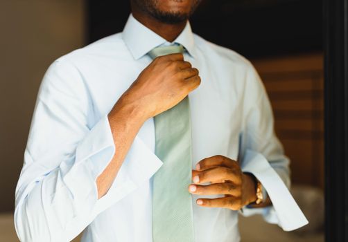 A man elegantly adjusting his tie, dressed smartly in a formal setting indoors.