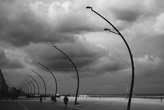 Monochrome view of a beach promenade with streetlamps under a cloudy sky.