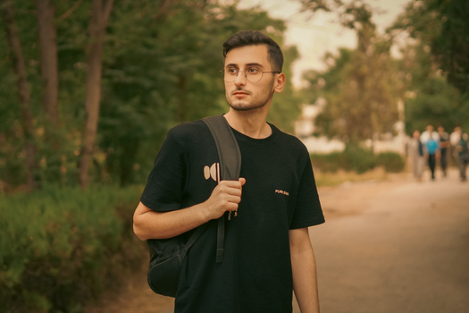 Portrait of a young man with glasses walking on a park path, carrying a backpack.