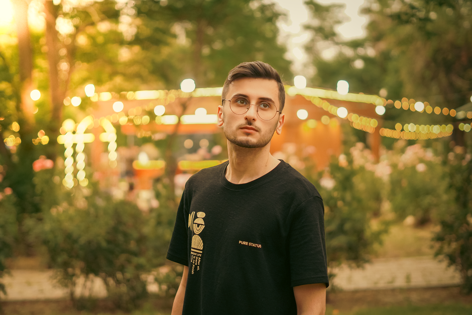 A stylish young man stands in a garden during twilight with festive string lights in the background.