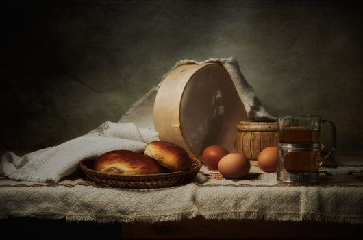 A rustic still life featuring freshly baked bread, eggs, and beer on a cloth-covered table.
