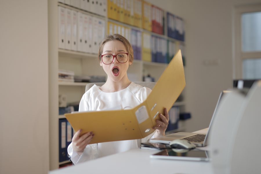 Surprised woman with eyeglasses reading a file in an office filled with shelves of records.