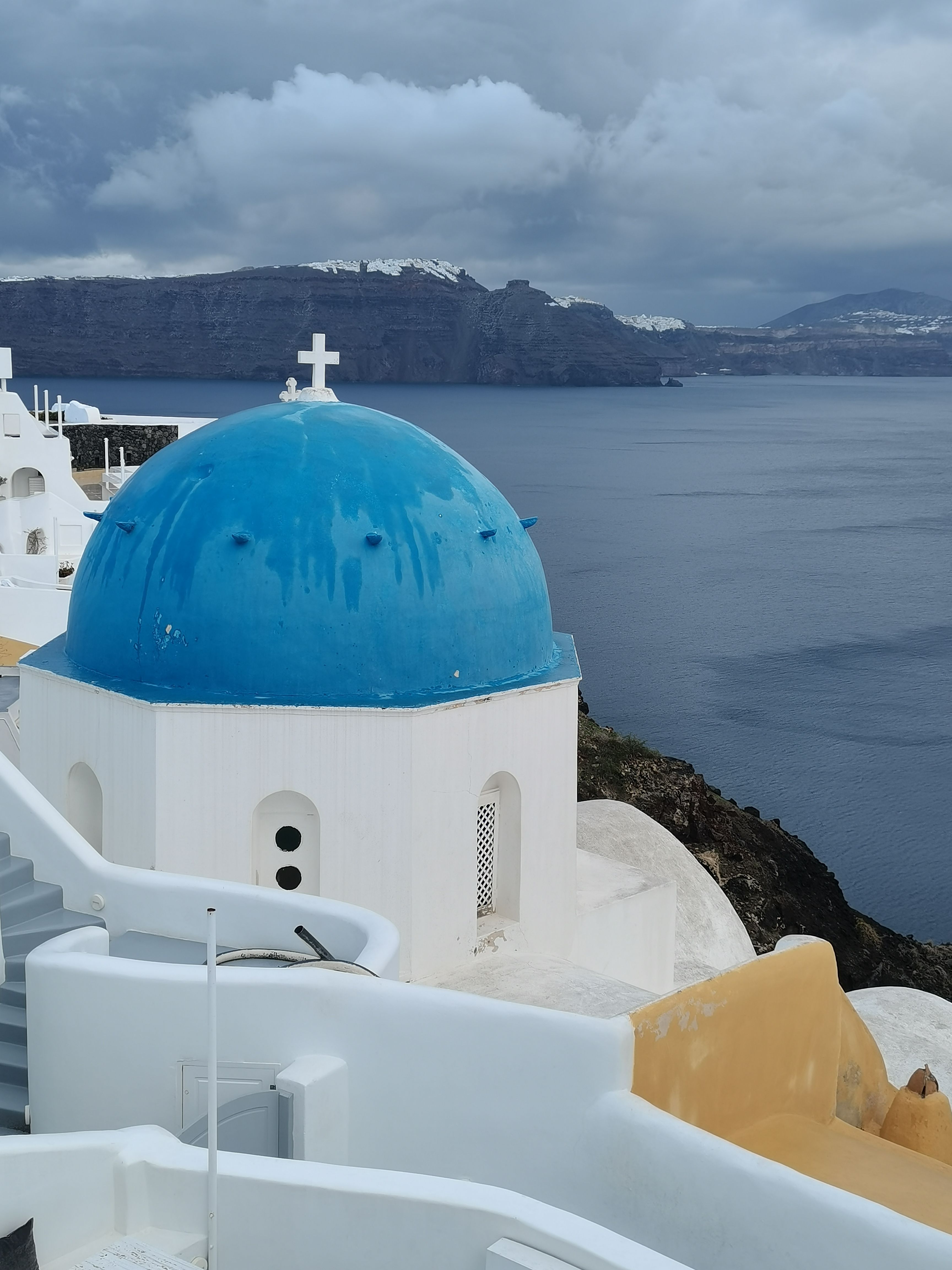 Iconic white and blue dome church in Santorini with a view of the Aegean Sea under a cloudy sky.