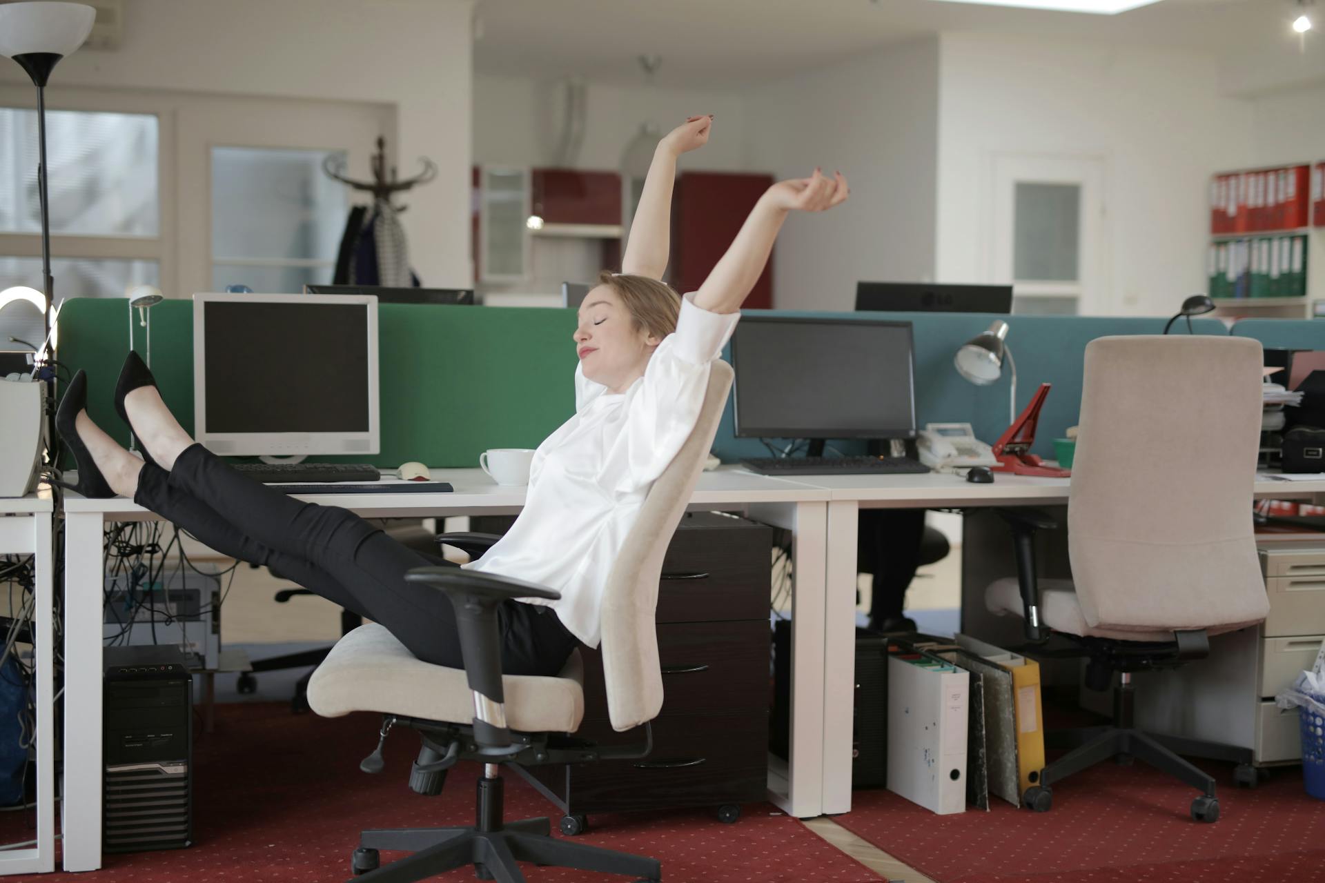 Zicht van de zijkant van vrolijke vrouwelijke werknemer in formele outfit zittend op een kantoor stoel met gekruiste benen op het bureau en rekken terwijl het rusten tijdens het werk met gesloten ogen