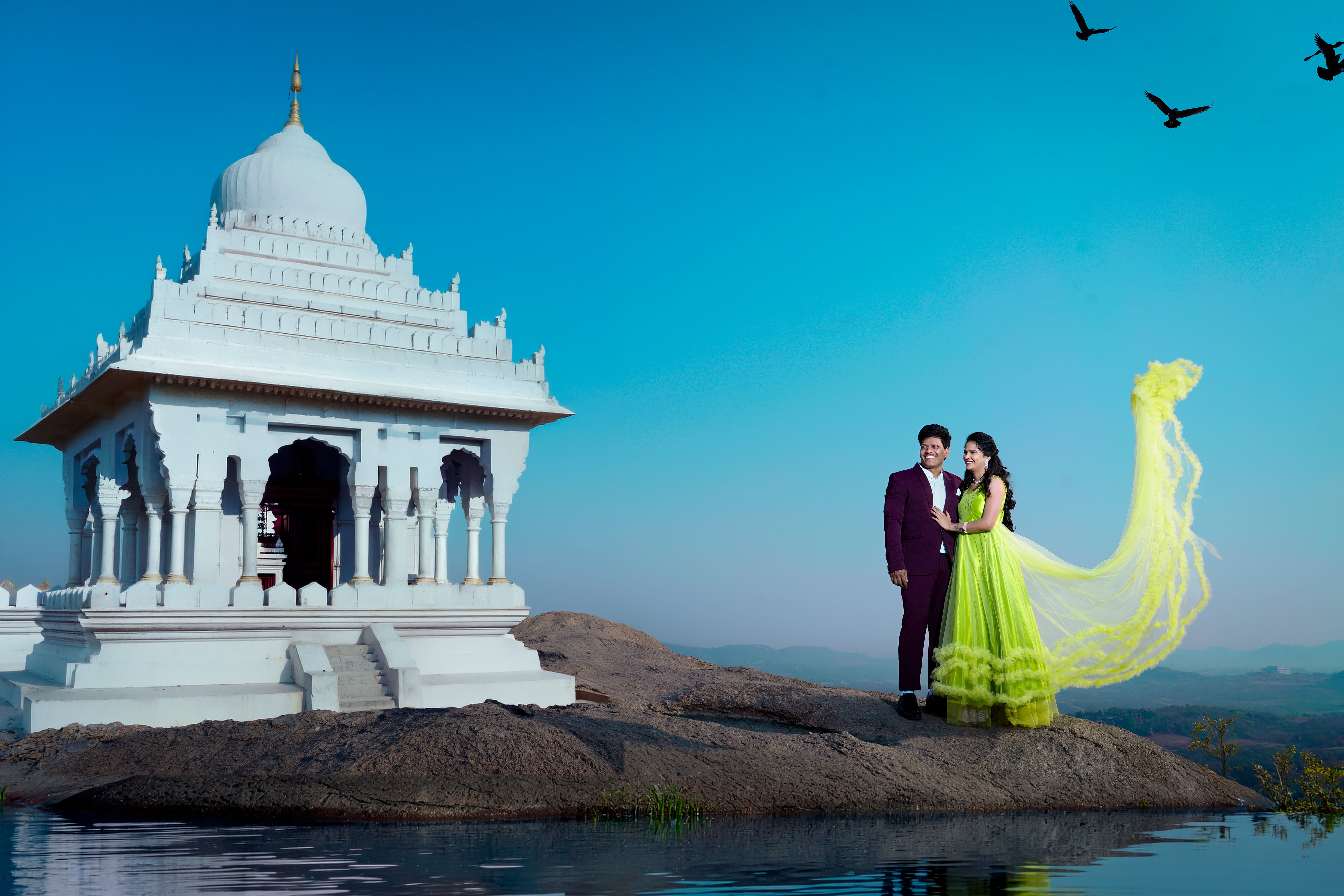 A couple in vibrant attire posing beside a white temple at sunset, reflecting in calm water.