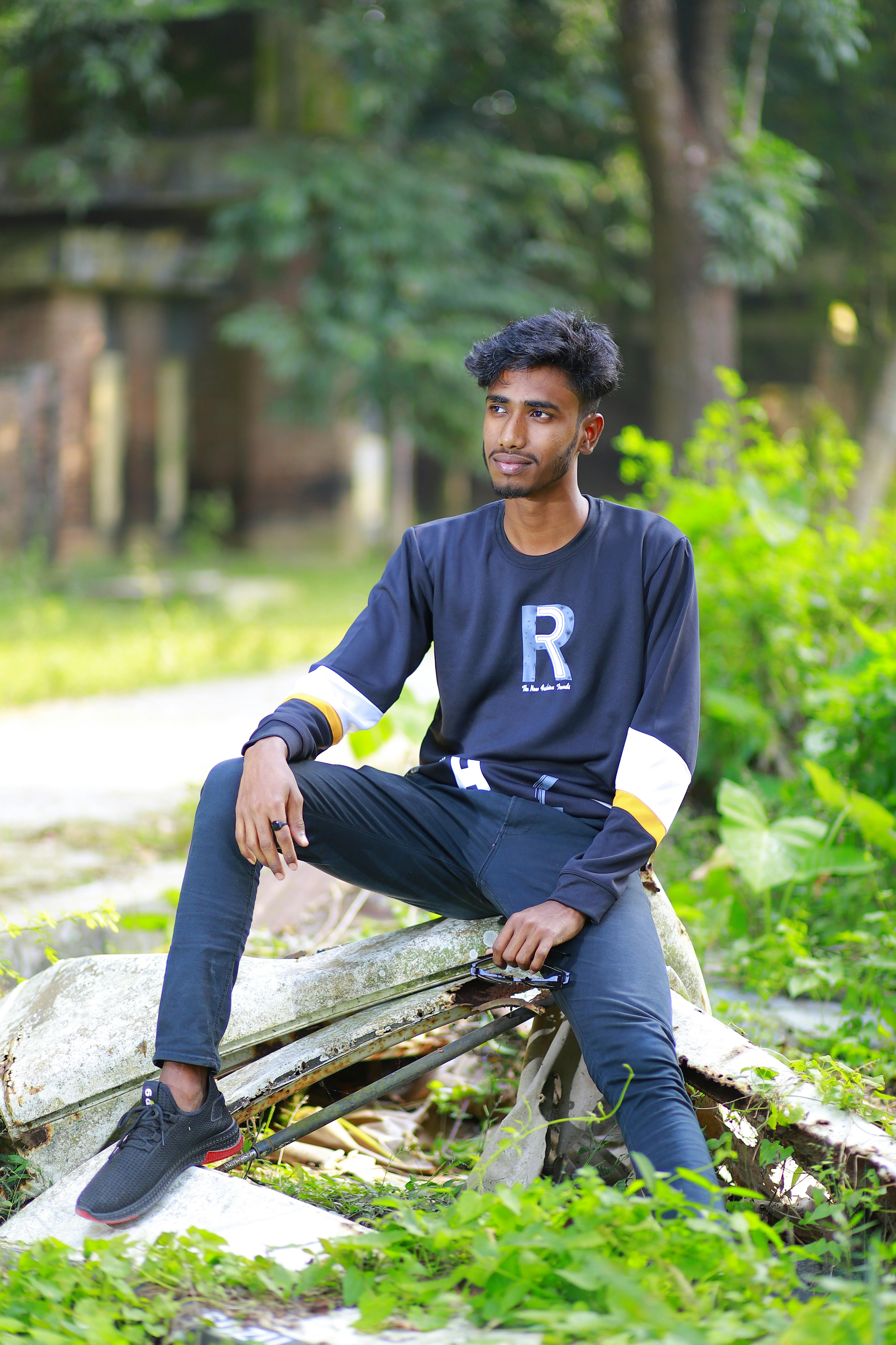 A young man sits casually near old concrete structures in a leafy outdoor setting.