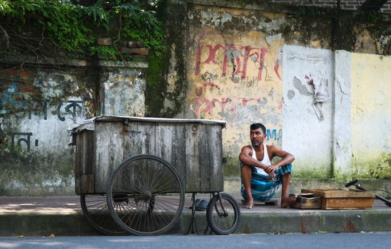 A street vendor takes a break beside his wooden cart against a rustic wall in Bangladesh.