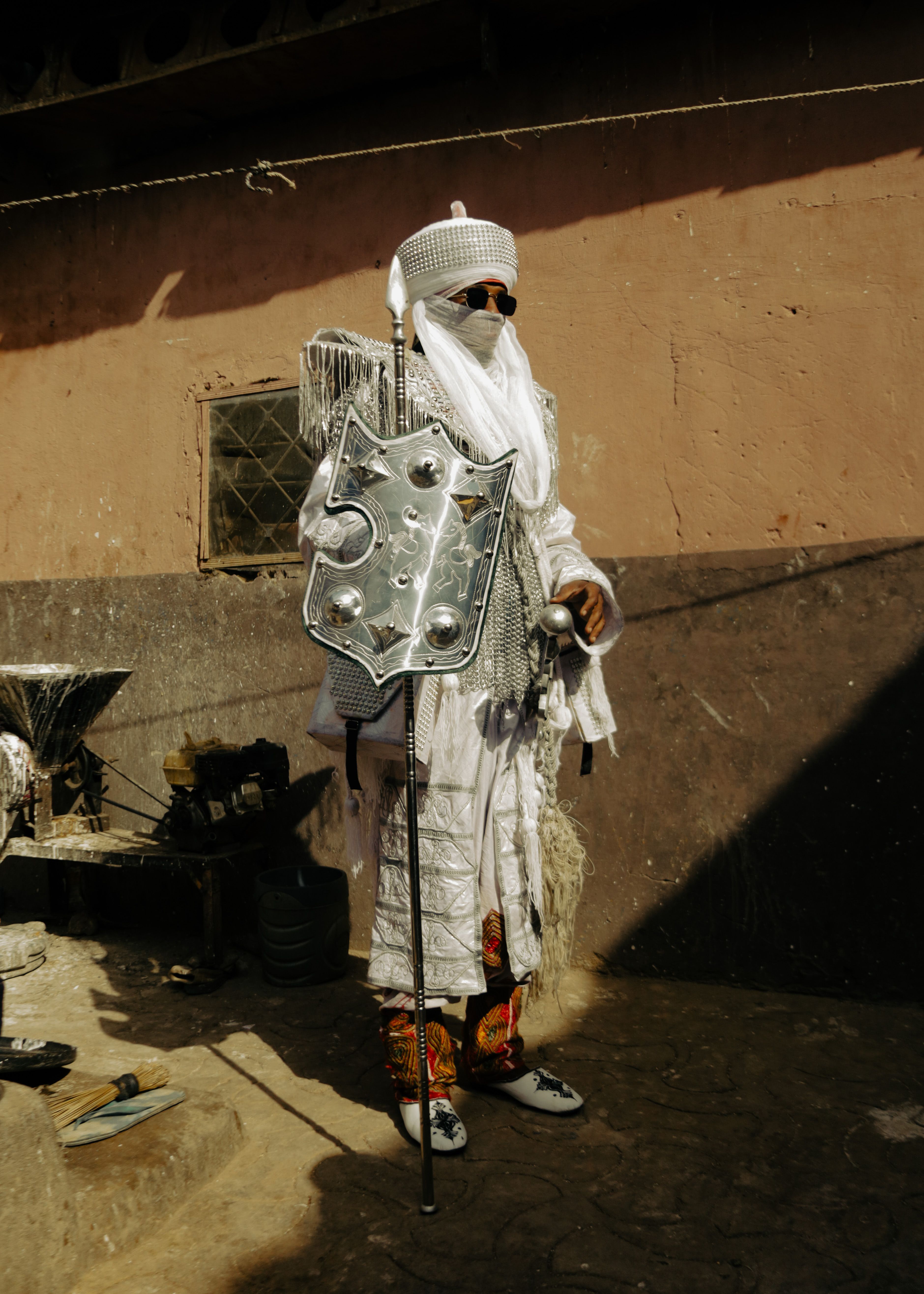 Man in traditional African warrior attire with intricate metallic shield outdoors.