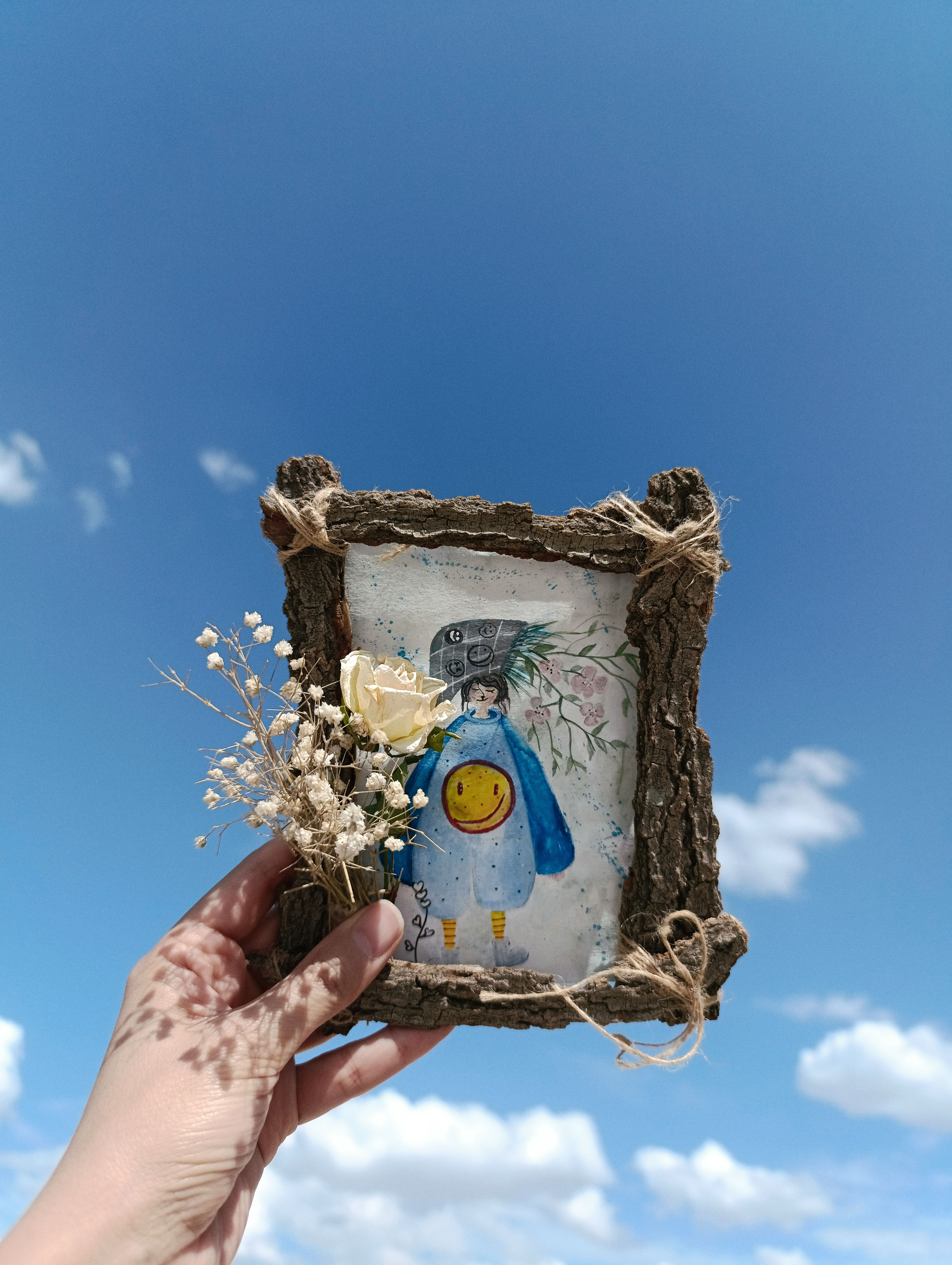 Rustic wooden frame with artwork and flowers held against a clear blue sky.