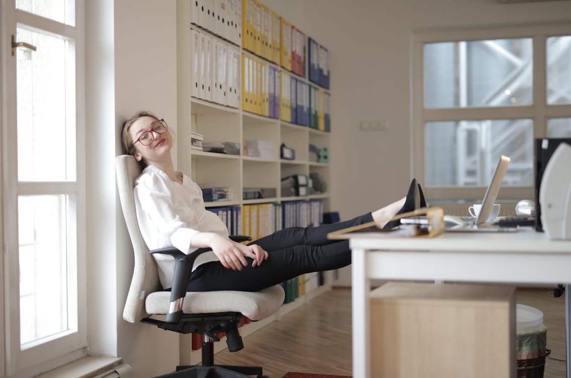 Free Side view of content female employee wearing formal clothes and eyeglasses sitting on chair with crossed feet on table and chilling during work with closed eyes in modern office Stock Photo