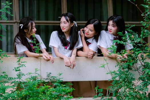 Four teenage friends in school uniforms chatting on a balcony surrounded by greenery.