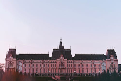 Exterior of aged masonry building with pink walls and forged peaks on roof with rectangular and arched windows near trees under serene sky