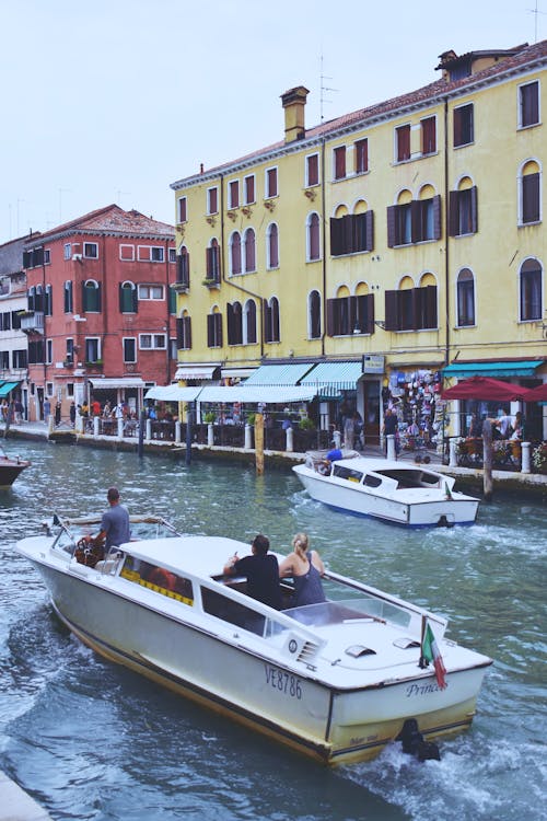 From above back view of anonymous travelers on motorized boat with boatman sailing on rippled water canal while exploring city and passing by facades of old houses during vacation